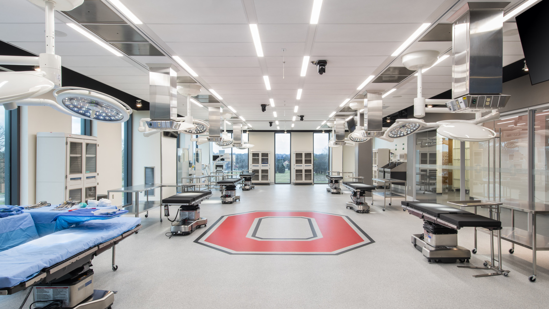 Inside view of room with 6 hospital beds with 2 circular lights hung above, tables next to the bed and red O on floor