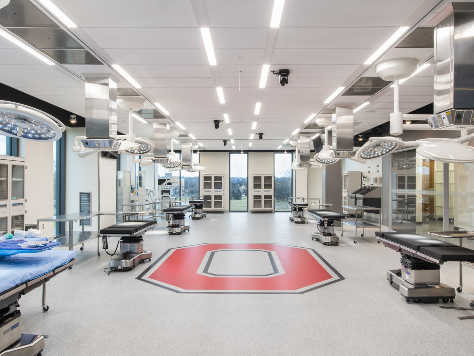 Inside view of room with 6 hospital beds with 2 circular lights hung above, tables next to the bed and red O on floor