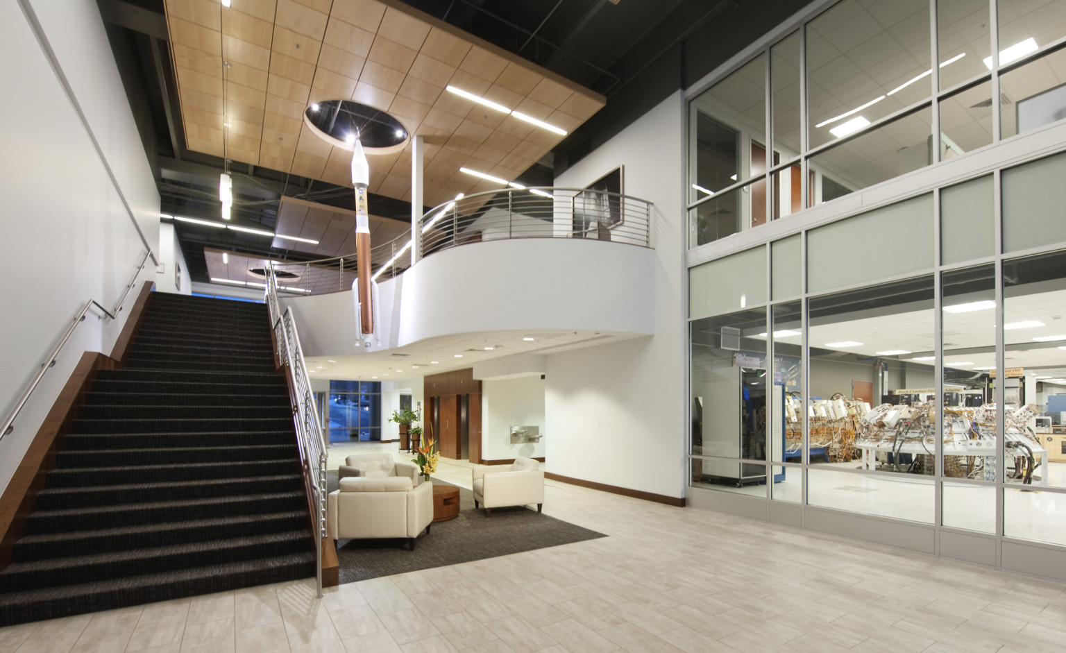 Double height atrium with wood panel drop ceiling detail over white arm chairs. Right, interior windows look into labs