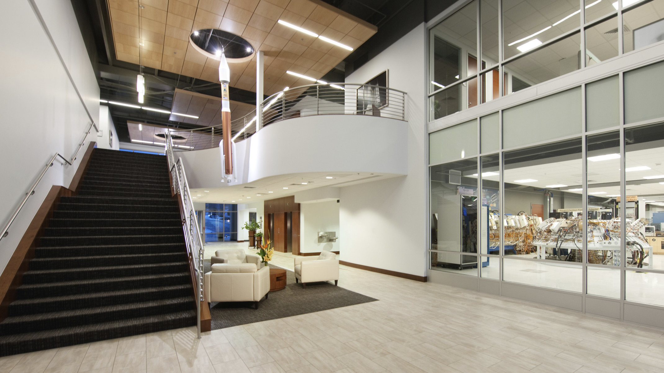 Double height atrium with wood panel drop ceiling detail over white arm chairs. Right, interior windows look into labs