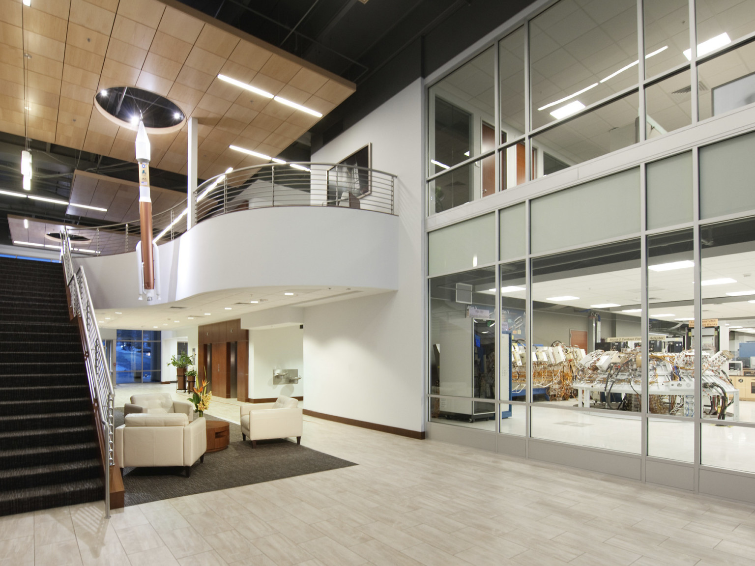 Double height atrium with wood panel drop ceiling detail over white arm chairs. Right, interior windows look into labs