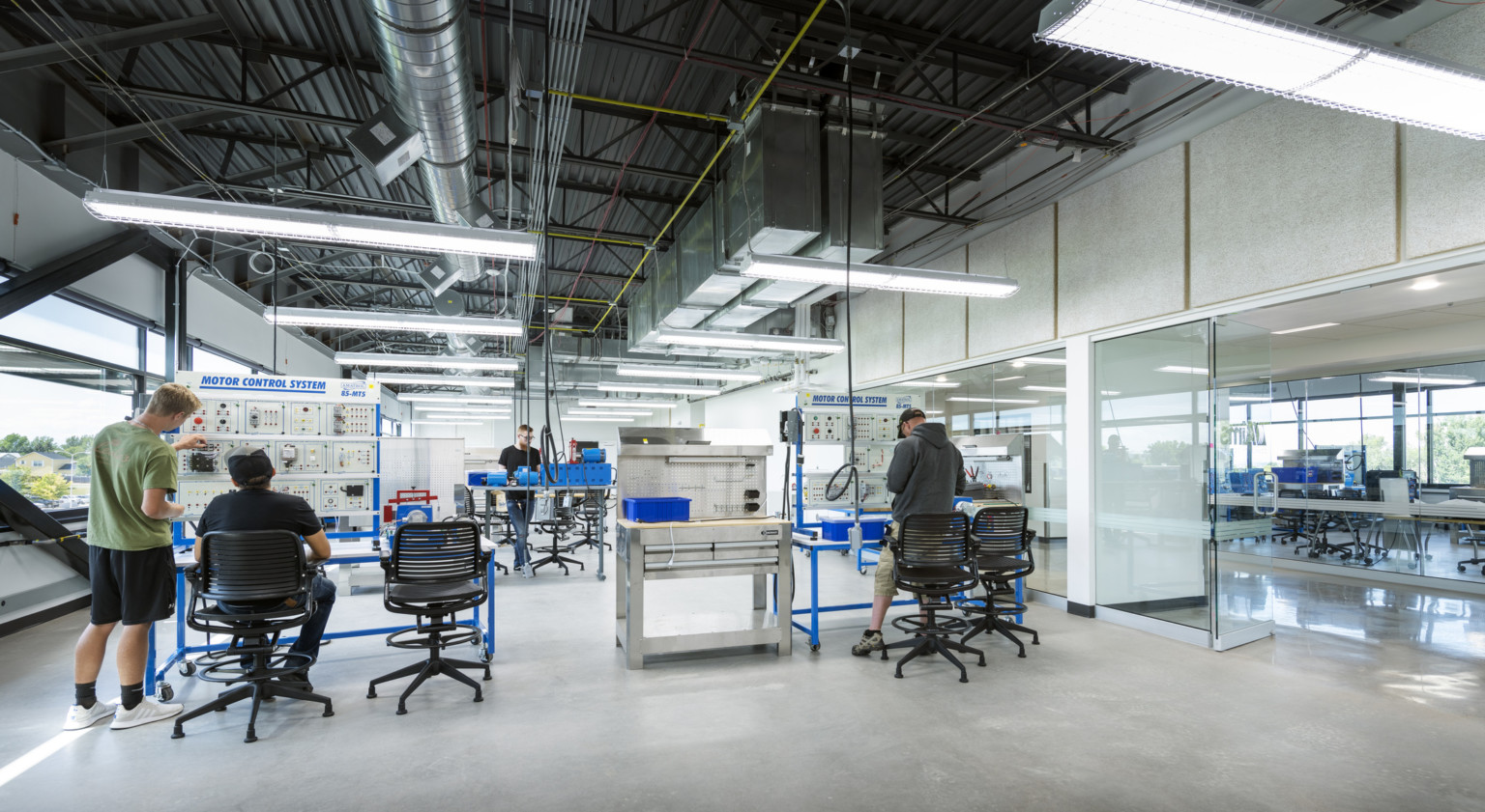 People work at Motor Control Systems in glass walled lab with exterior windows, left. Exposed ducts and hanging lights