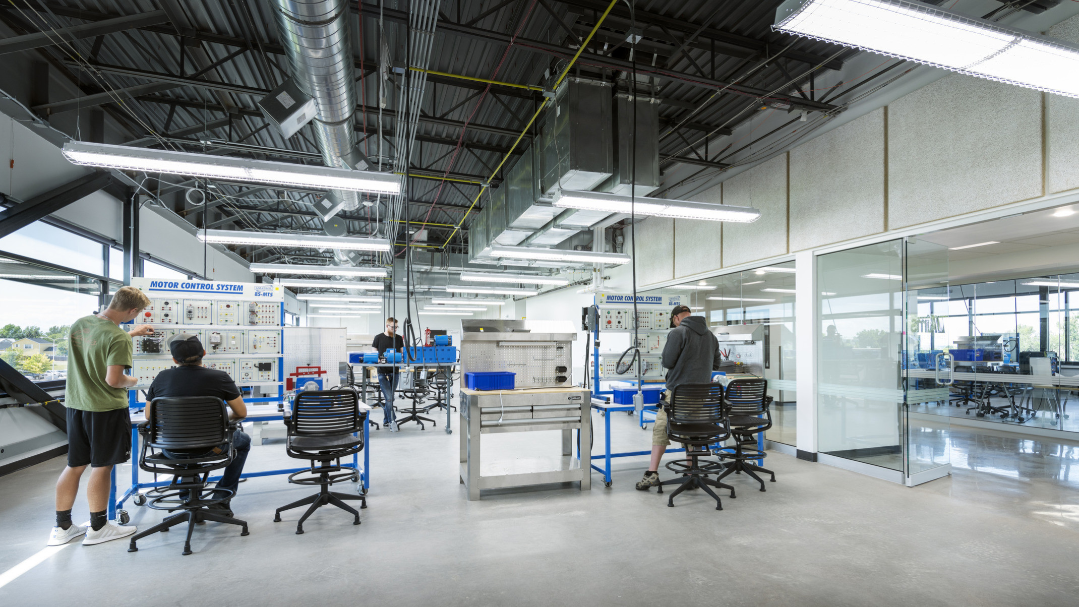 People work at Motor Control Systems in glass walled lab with exterior windows, left. Exposed ducts and hanging lights