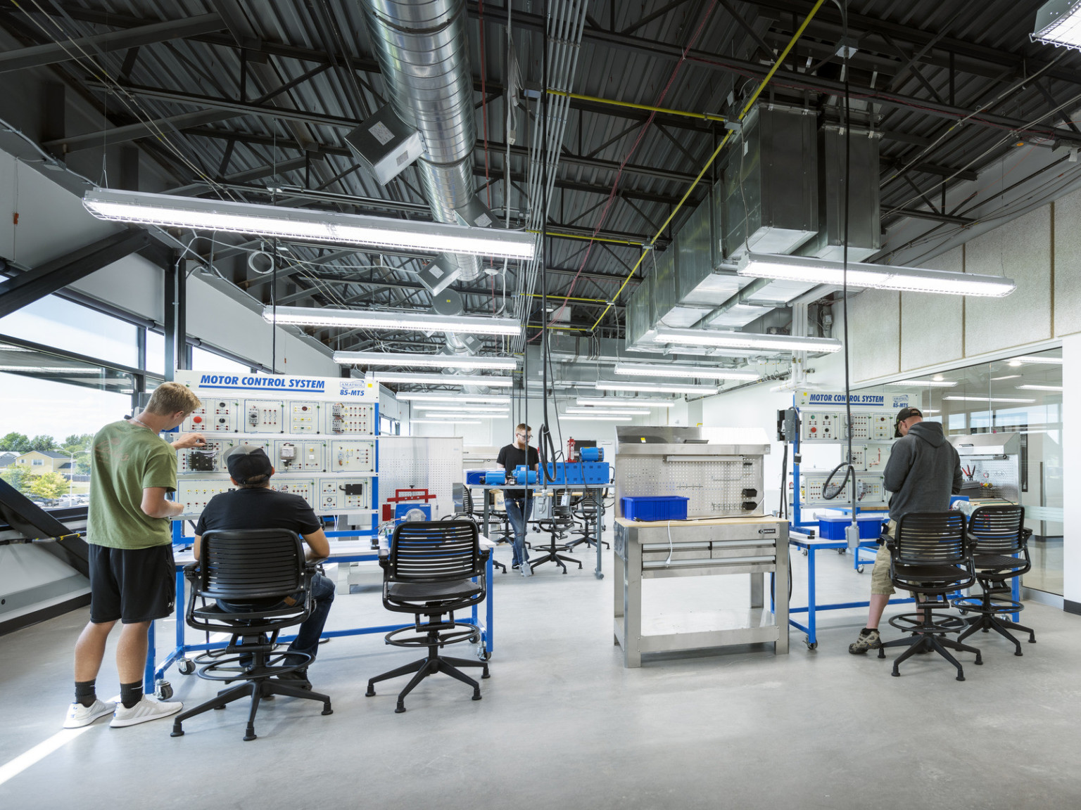 People work at Motor Control Systems in glass walled lab with exterior windows, left. Exposed ducts and hanging lights