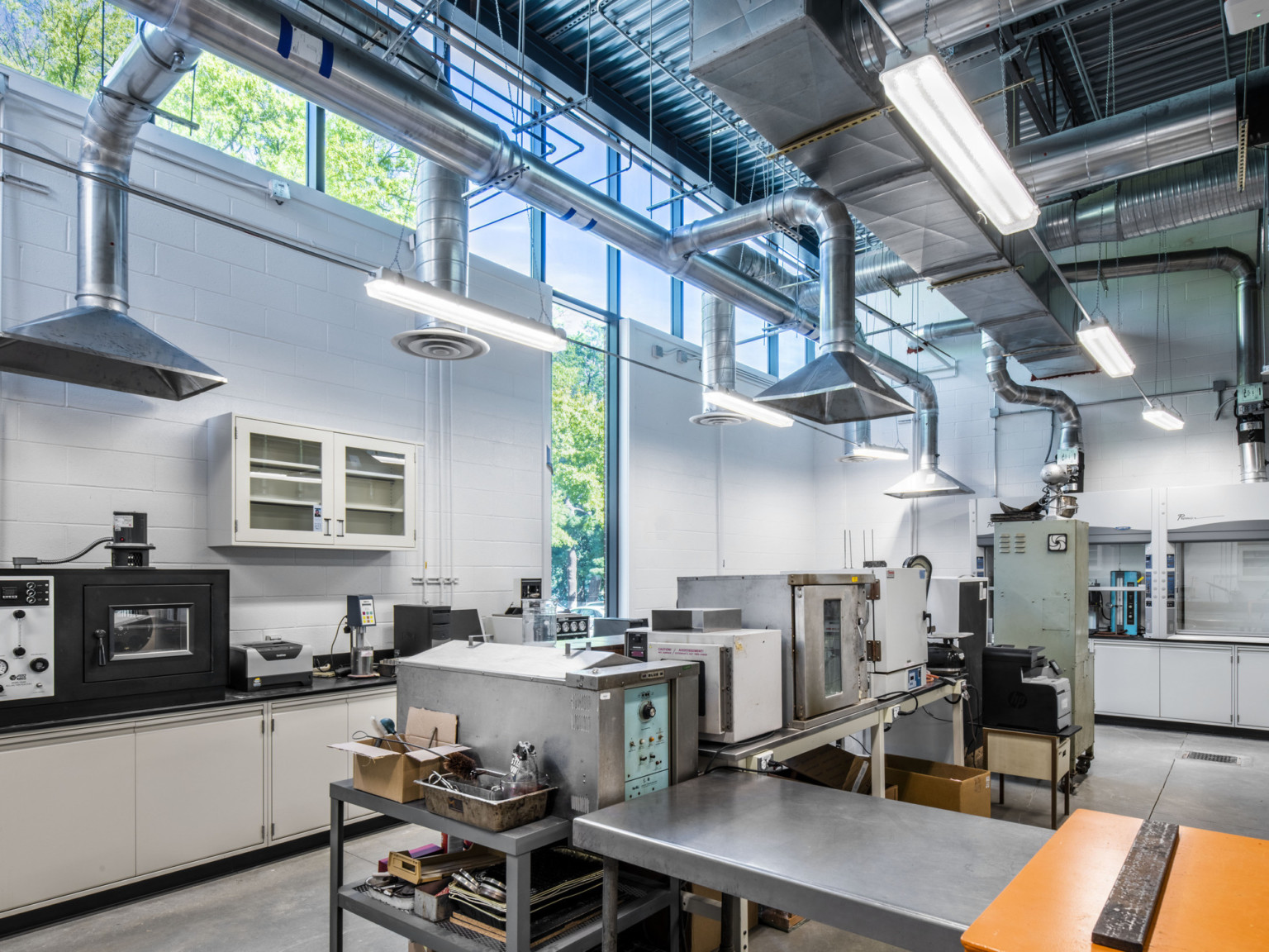 Double height white cement block lab with exposed ducts above lab equipment. View, left, through windows to green space