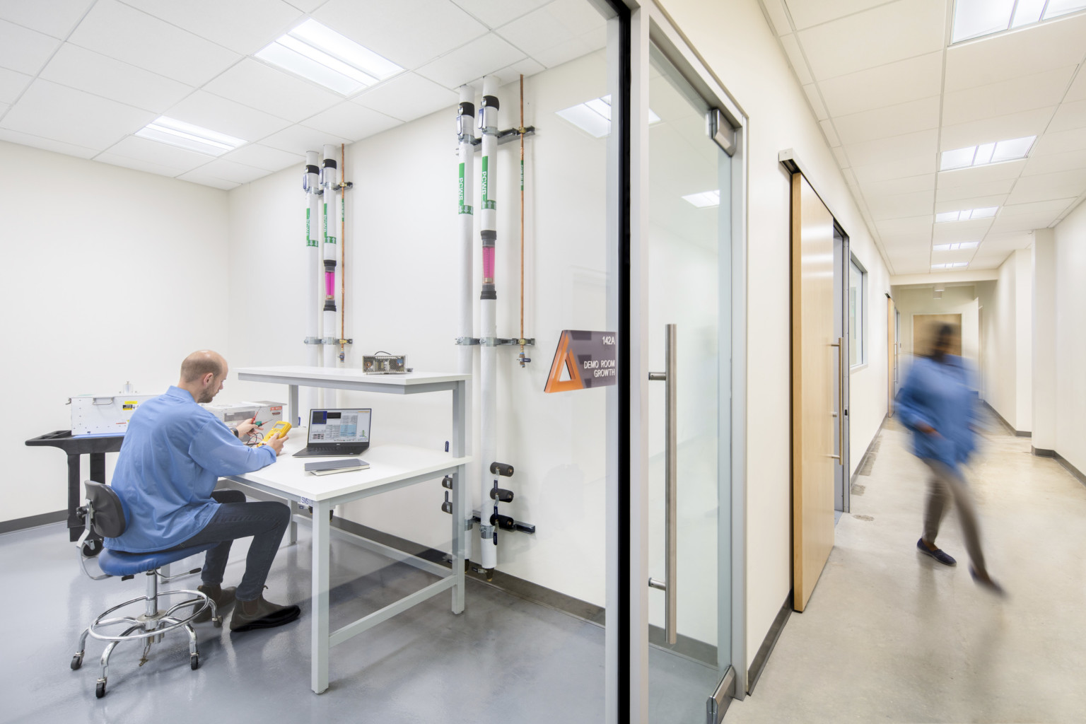 a glass room off a white hallway contains multilevel workspace and tubes connecting to wall. A man works with equipment