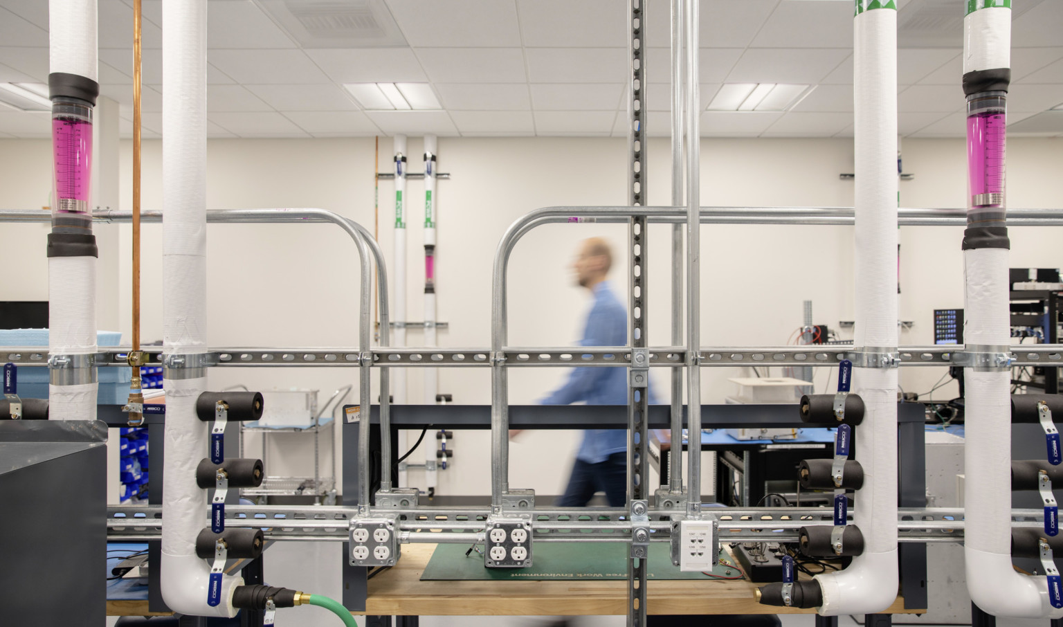 lab with white parallel vertical tubes with pink centers and electric piping bisecting horizontal and vertical metal tracks.