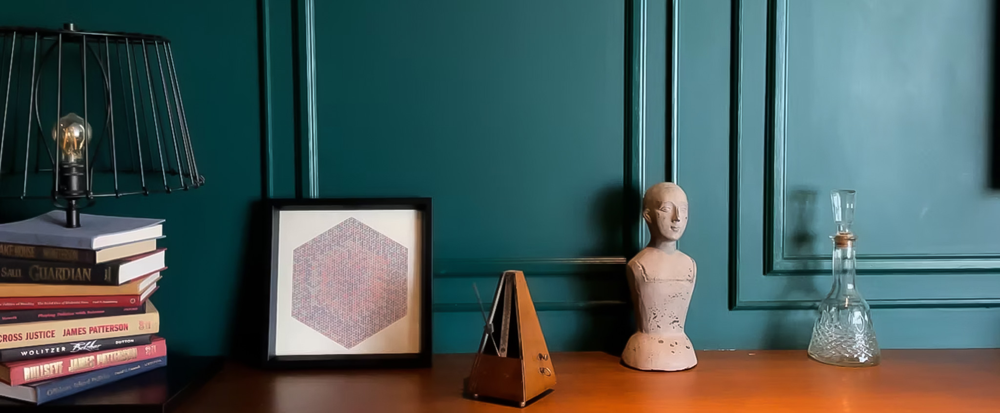 dark green wall with molding details behind a lamp made from stack of books, framed art print vintage metronome and cast bust