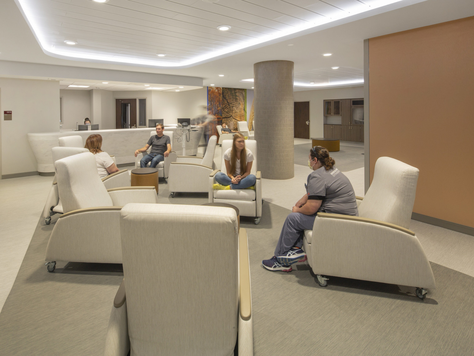 People in wheeled arm chairs in groups around round tables in big room with illuminated recessed ceiling detail, orange wall