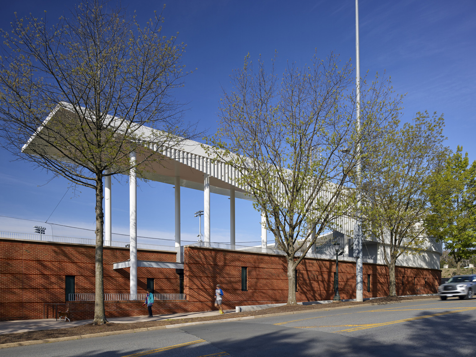 Exterior of back of stadium. Trees by brick wall with thin windows built into hill viewed from across the street. White canopy