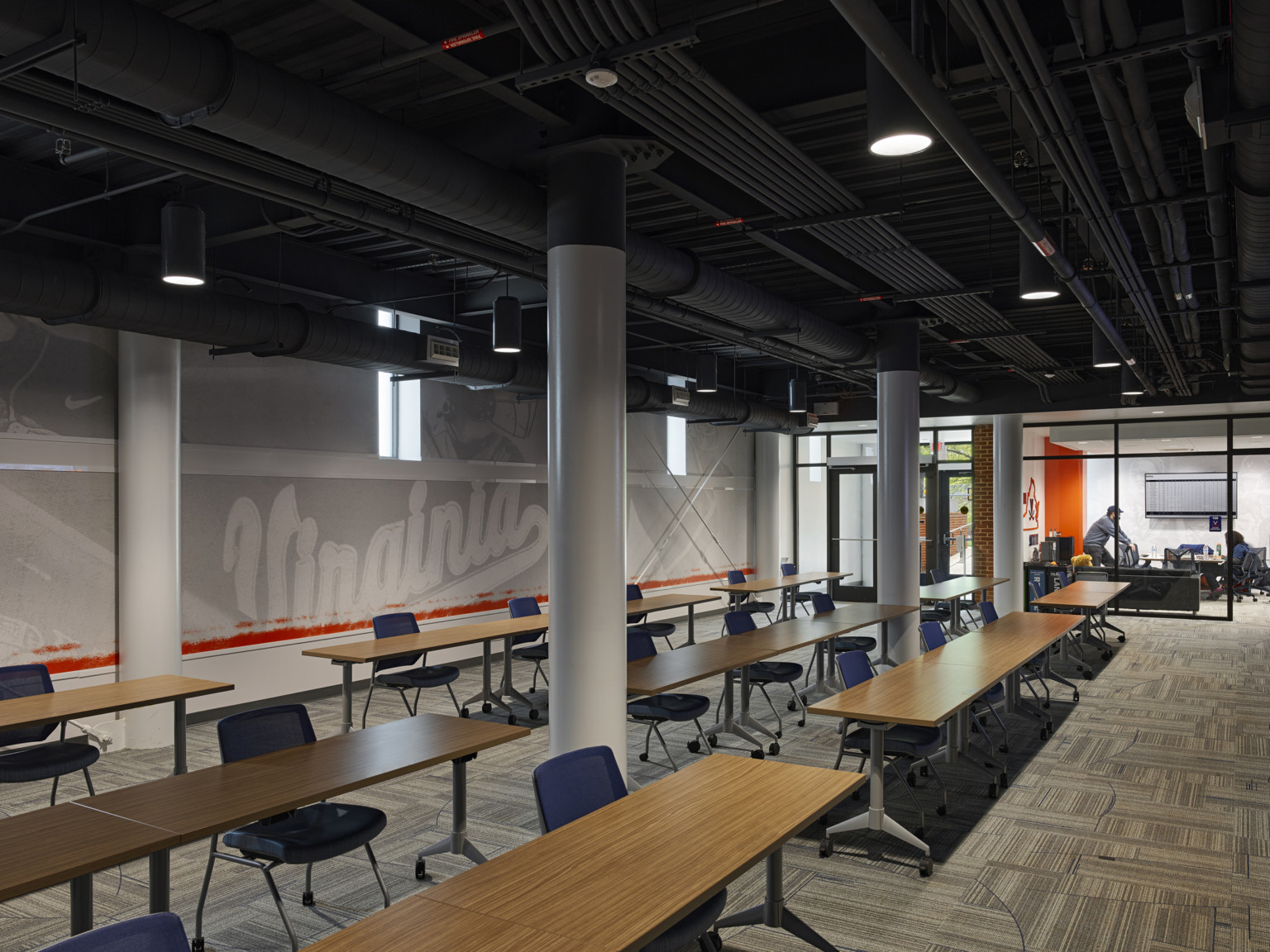 Interior, black and white mural behind rows of wood tables with rolling chairs. White columns, black ceiling, exposed ducts