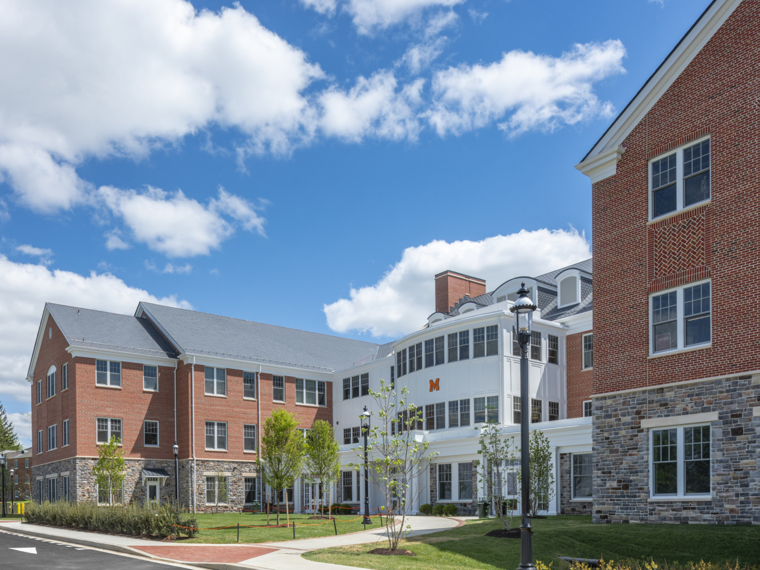 three-story building with stone 1st floor and brick upper connect to white addition at center with large windows and columns under blue skies with fluffy white clouds