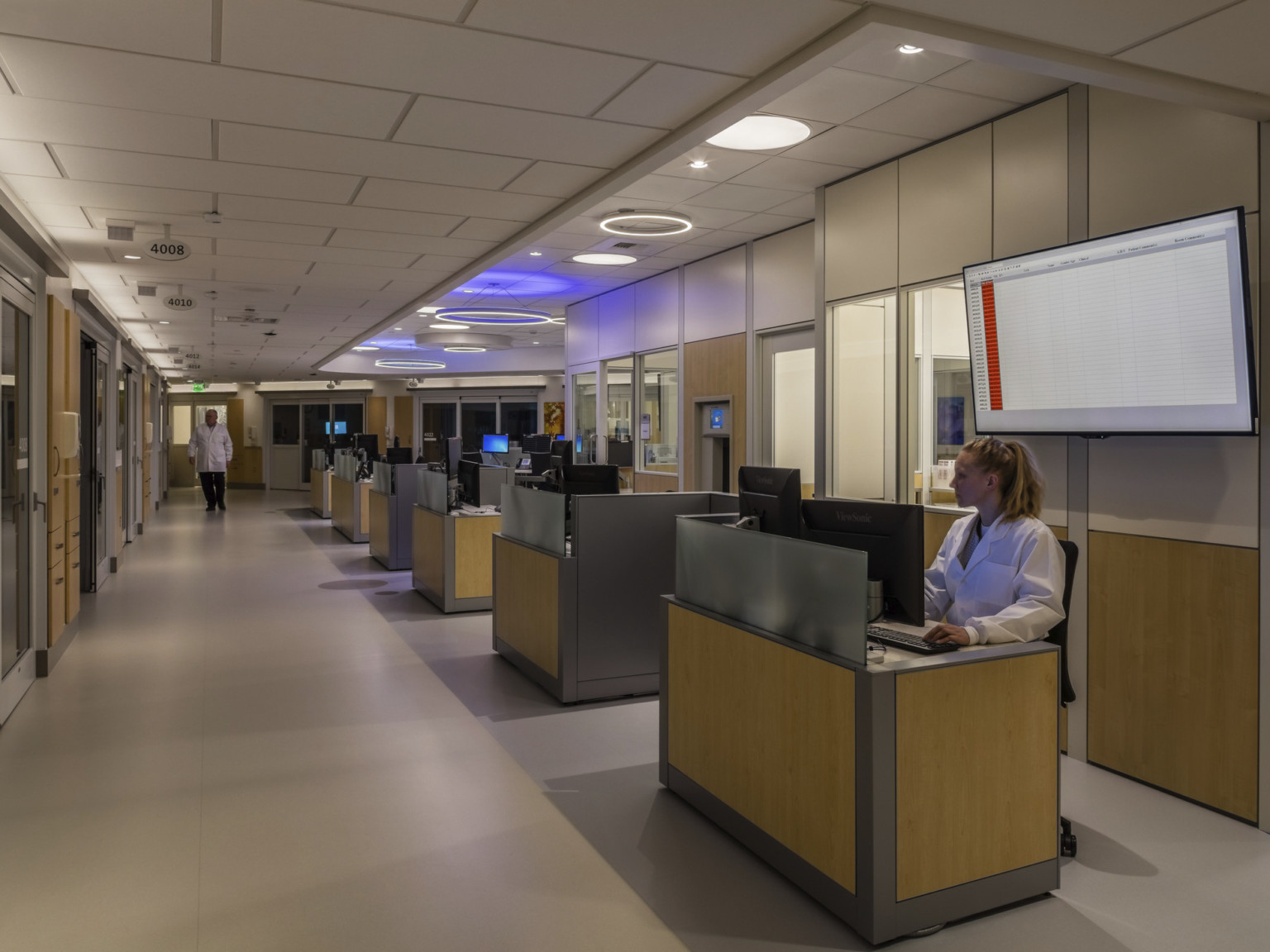 Individual work stations along wall of hallway with view to patient rooms under round accent lights in recessed ceiling area