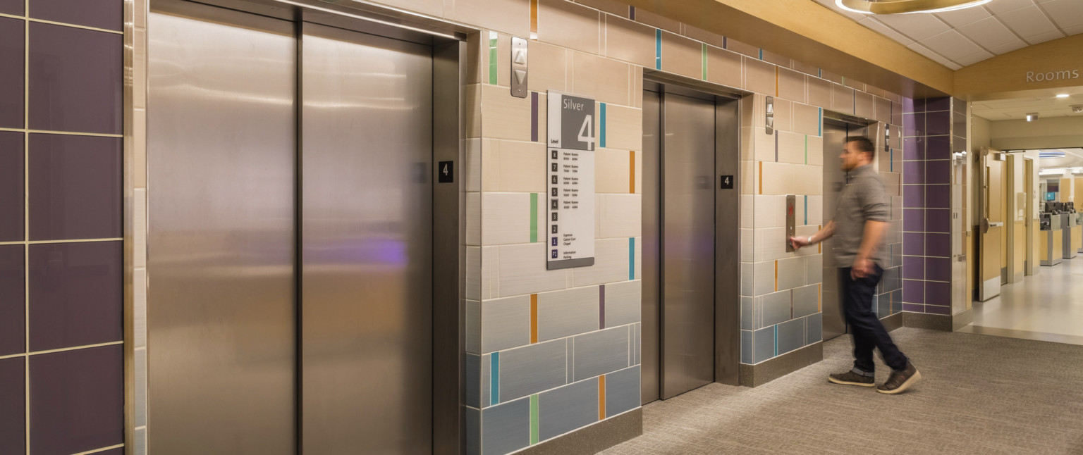 Elevator lobby with multicolor wall panels surrounding doors and purple tiles at end. circular light hangs overhead
