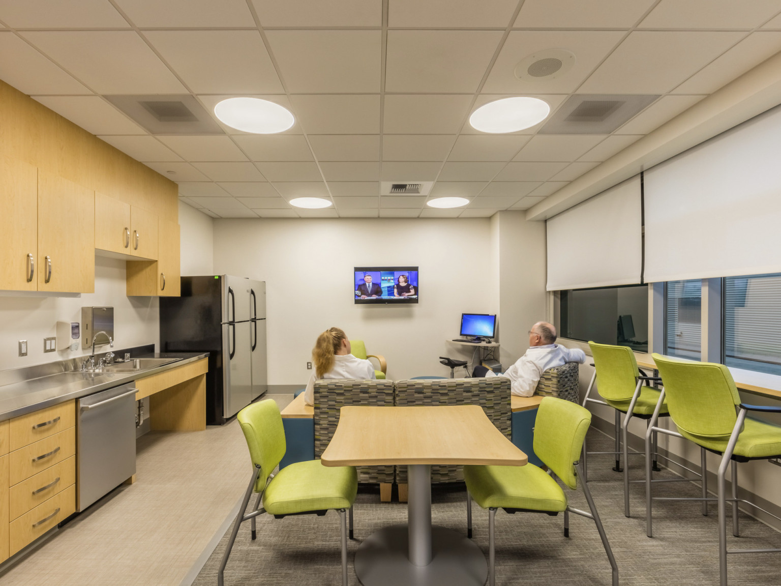 Comfortable seating in front of tv behind table and green chairs. Kitchenette to left in white room with wood cabinets