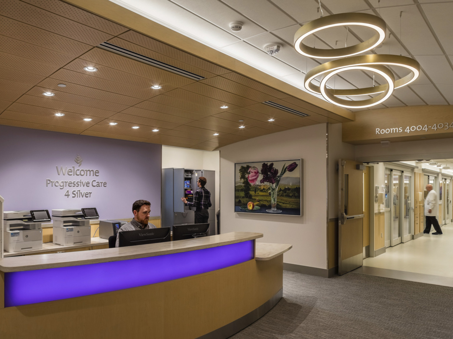 Reception desk with illuminated blue panel on front under modern round chandeliers in hallway. Office behind blue accent wall