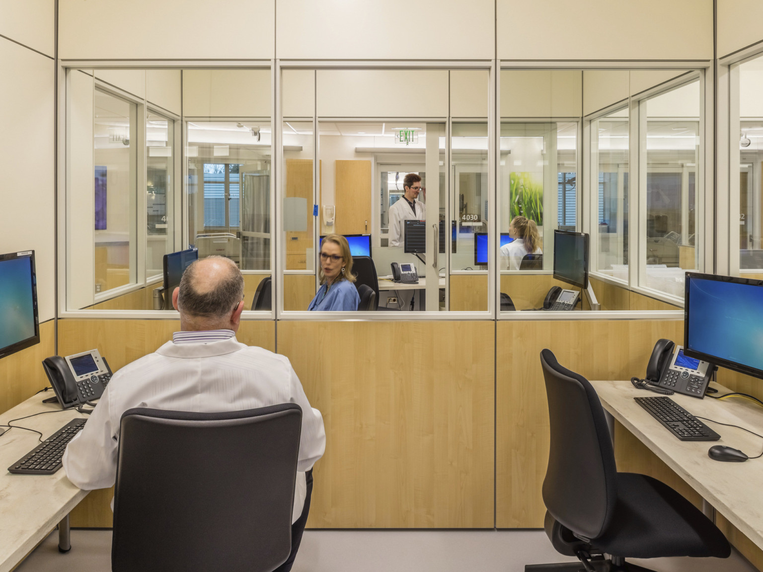 2 person work spaces with large windows above wood wall panels separating modern cubicles off hallway in healthcare facility