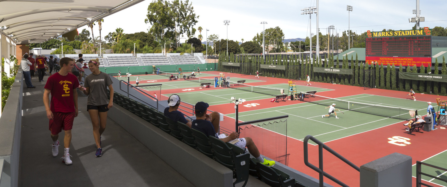 Covered walkway behind stadium seating looking to tennis courts. Tree lined far wall, scoreboard above with Marks Stadium sign