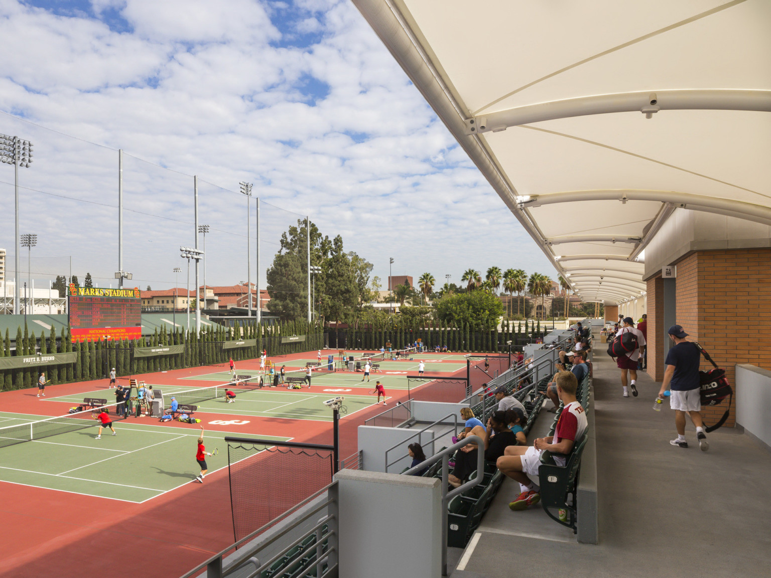 Covered walkway extends from brick building behind seating and tennis courts. Trees on left, far wall, Marks Stadium sign