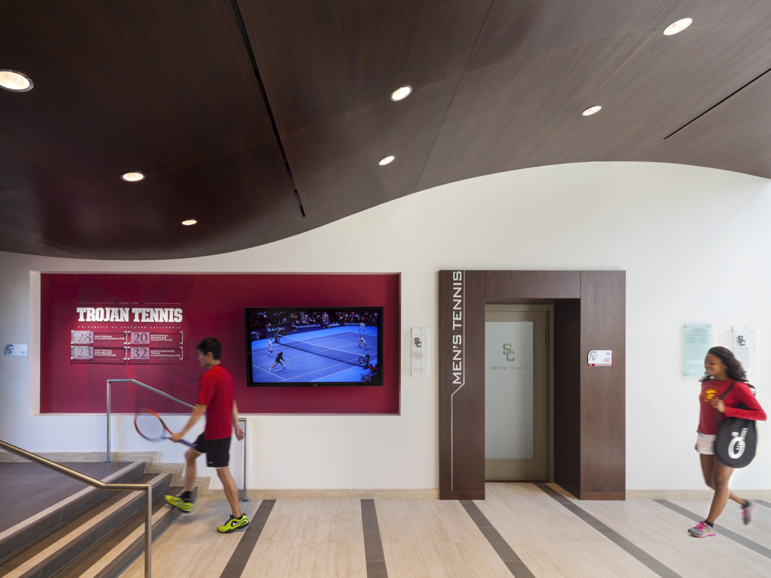 Curving wood ceiling over hallway. Red recessed wall detail with screen and Trojan Tennis sign by two tone steps