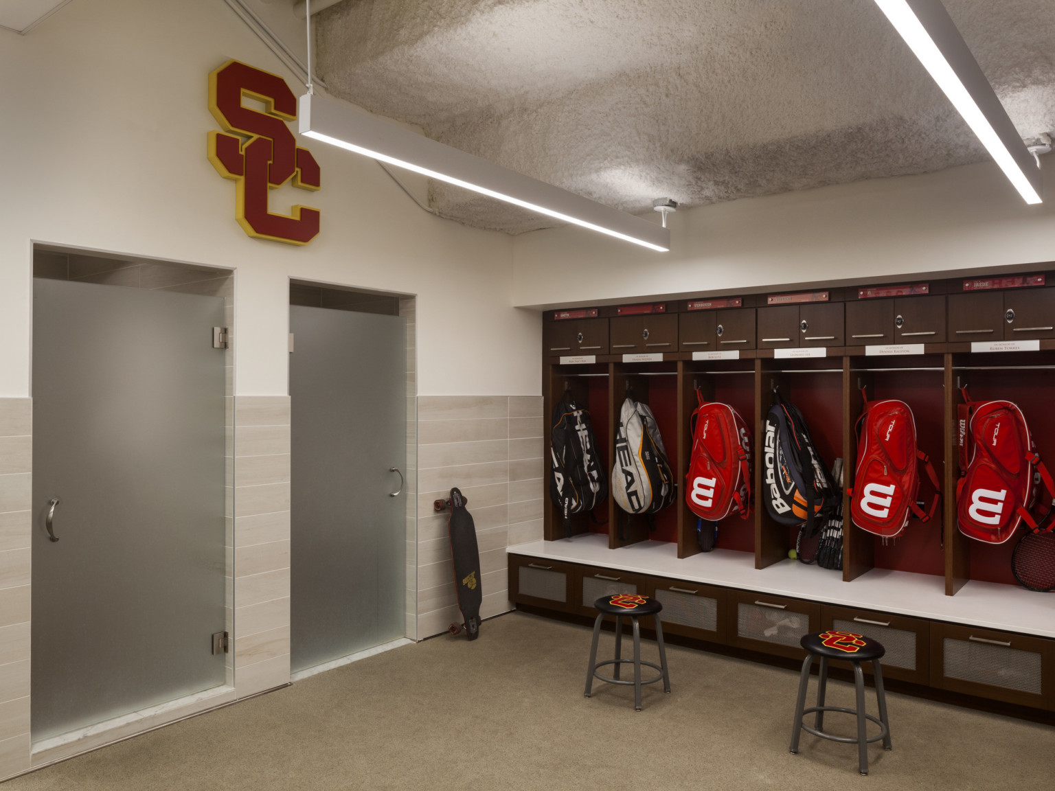 Locker room with textural stepped ceiling and hanging rod lights over wood lockers with white bench seat. Tiled showers left