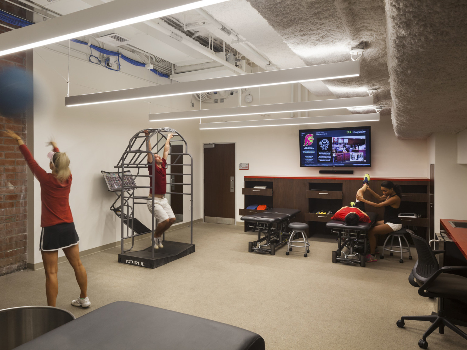 White exercise room with exposed brick and high stepped ceiling. Massage tables at back wall with wood cabinets of equipment