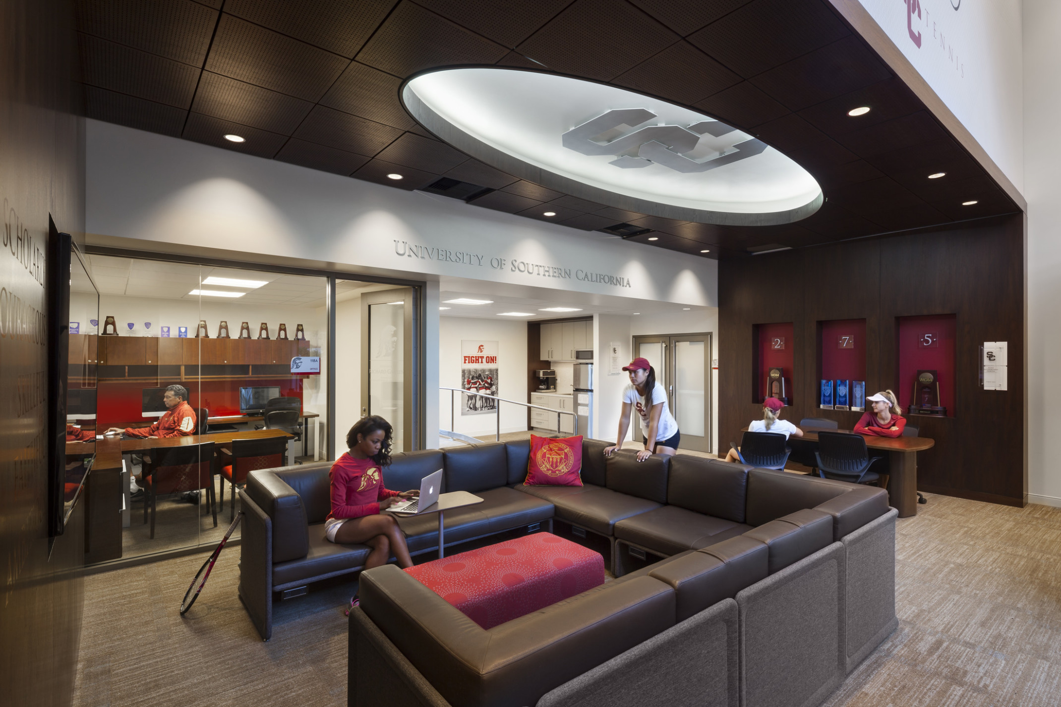 Brown sectional faces screen in common area. Kitchenette in back corner, glass walled office to left. Recessed ceiling detail