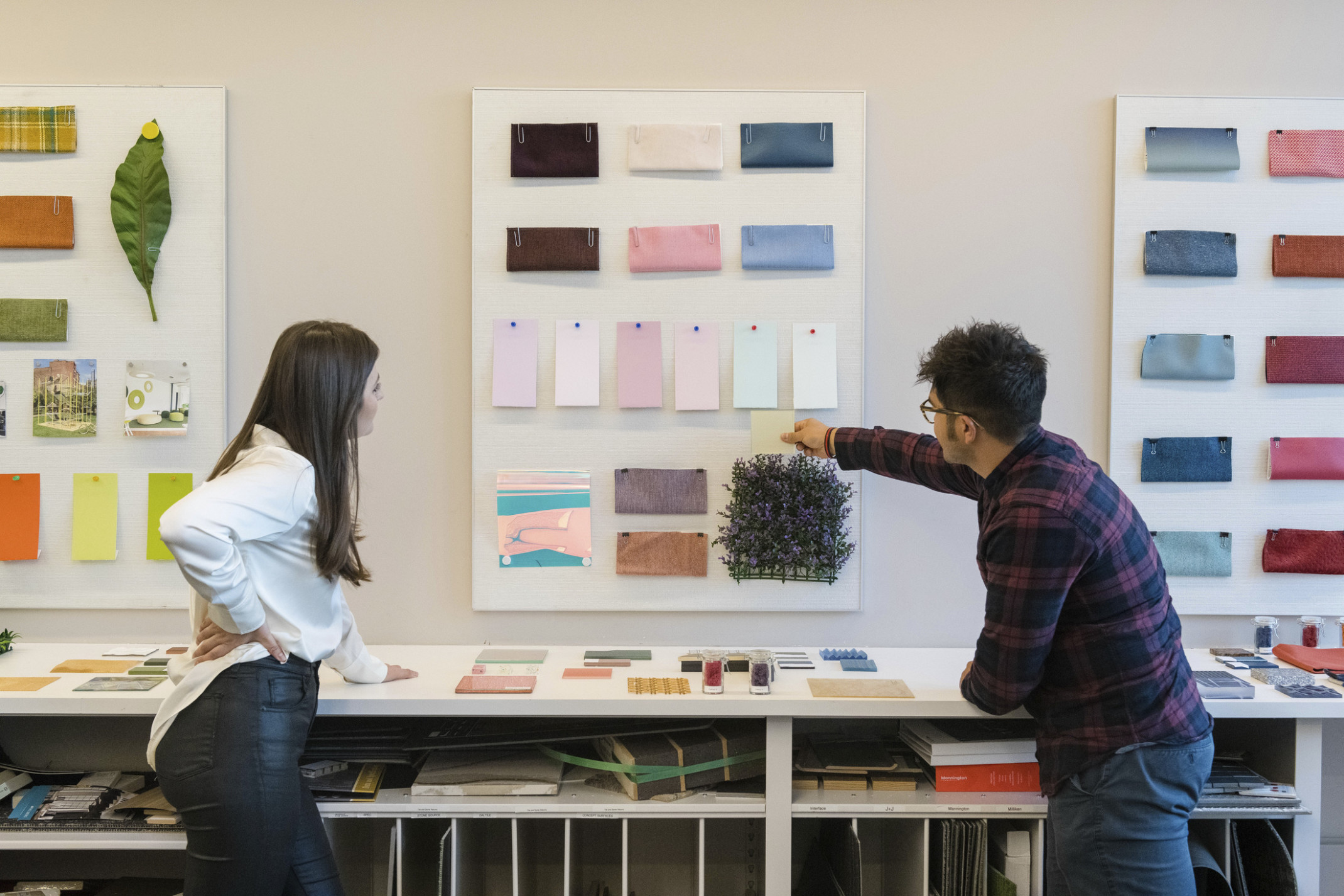 two people looking at a white board pinned on a wall filled with evenly spaced paper and fabric swatches in purple colorways