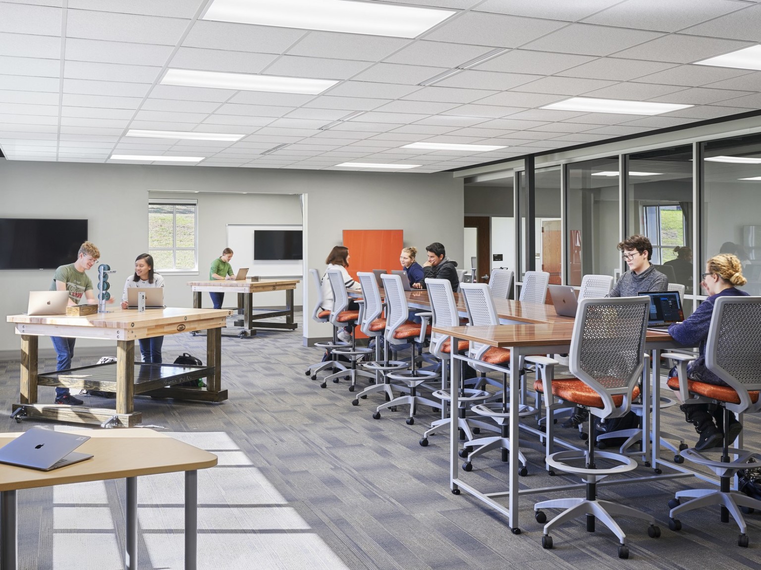 students working on an engineering project on the left with additional students collaborating at a long wood table on the right. Orange chairs, grey carpet.