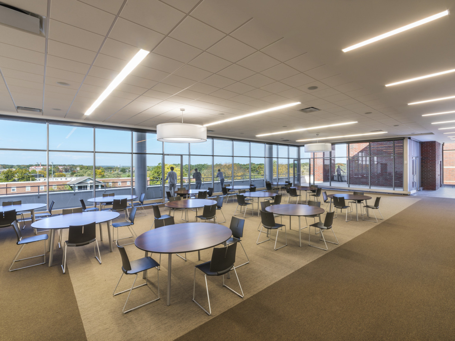 Interior flexible seating area with round tables. Floor to ceiling windows look to exterior balcony walkway overlooking campus