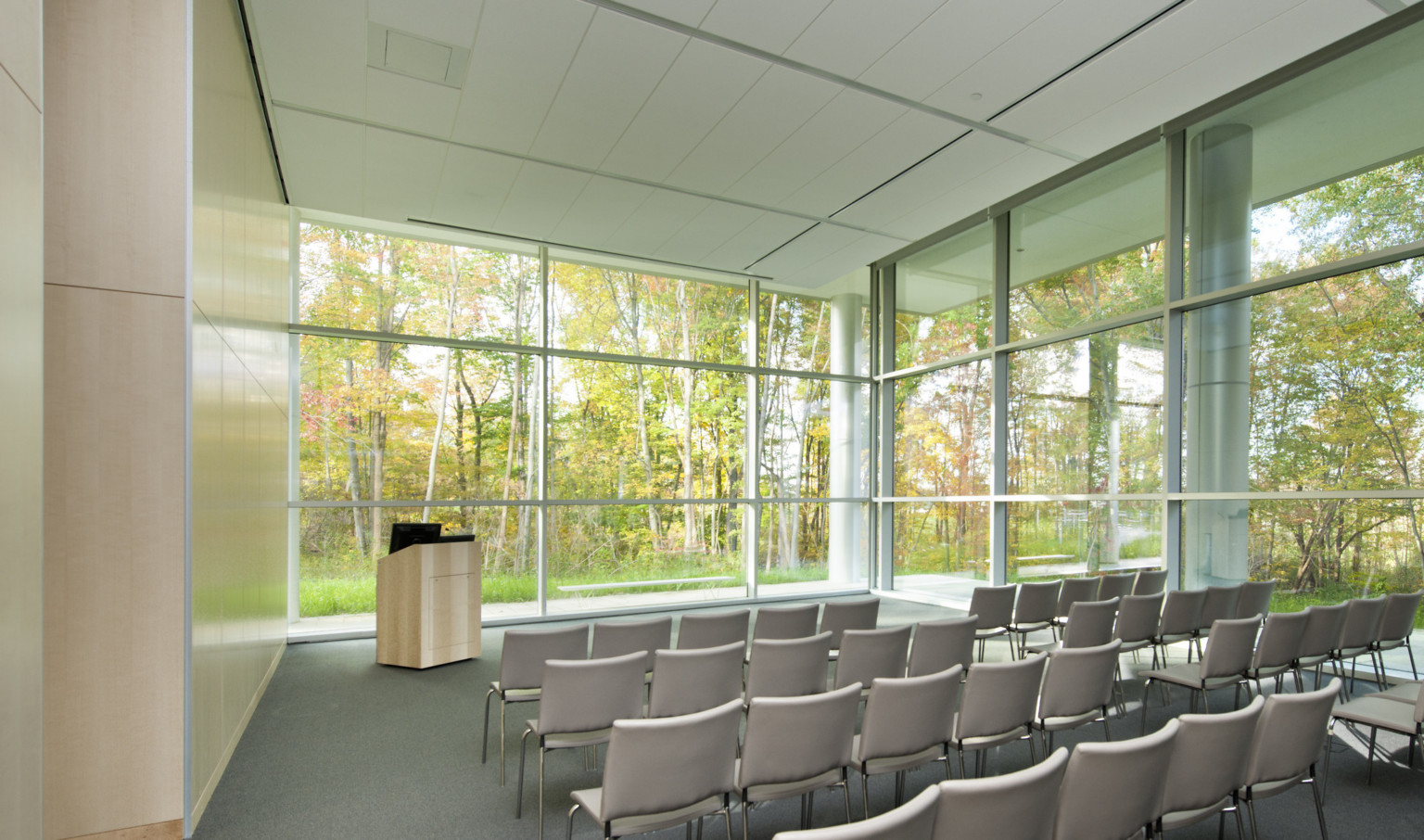 Auditorium with grey floor, wood left wall, and floor to ceiling windows on two walls with ground level view of trees.
