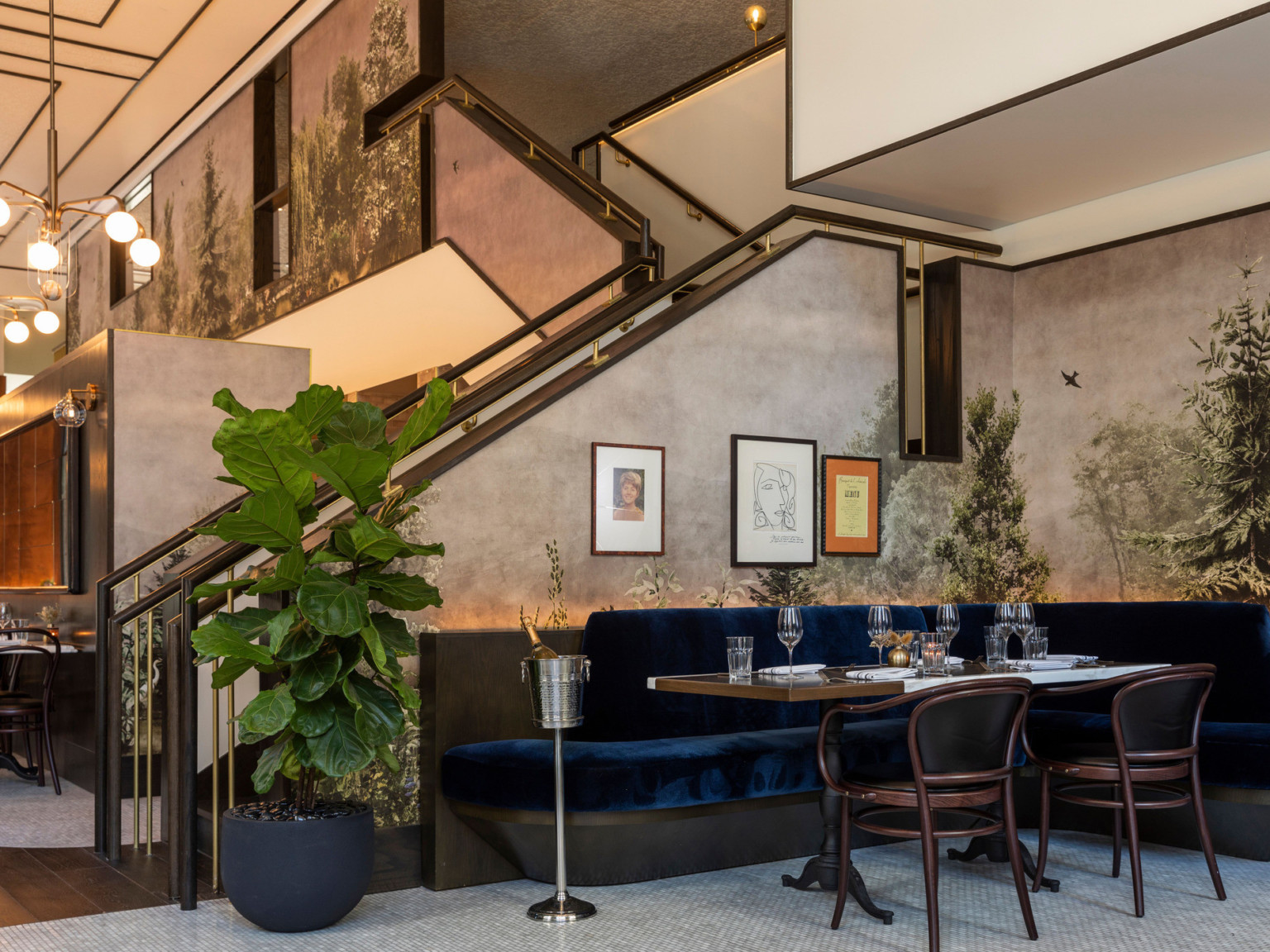 Blue velvet corner booth seating in front of snowy tree mural and stairs done to restaurant level. Modern chandeliers