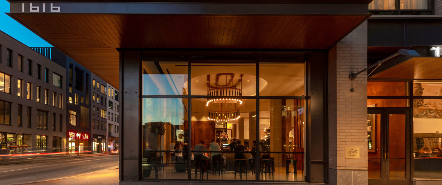 View through floor to ceiling window to street level lounge. Black canopy extends over sidewalk, wrapping around corner