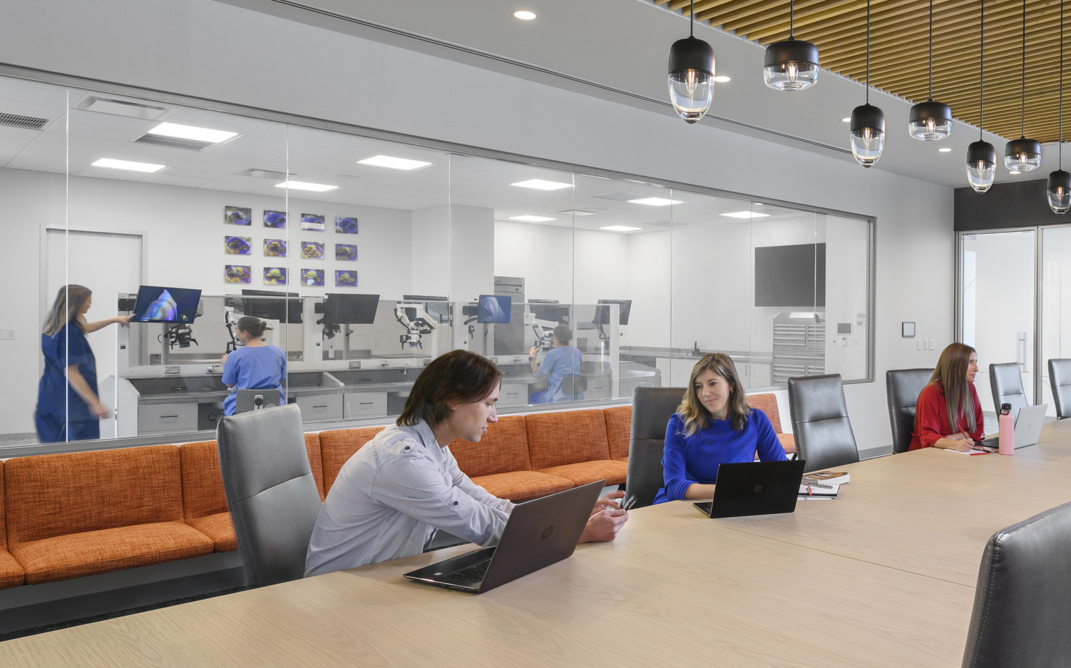 Conference room with wood slat ceiling over long table with pendant lights down center. Black back wall with large screen