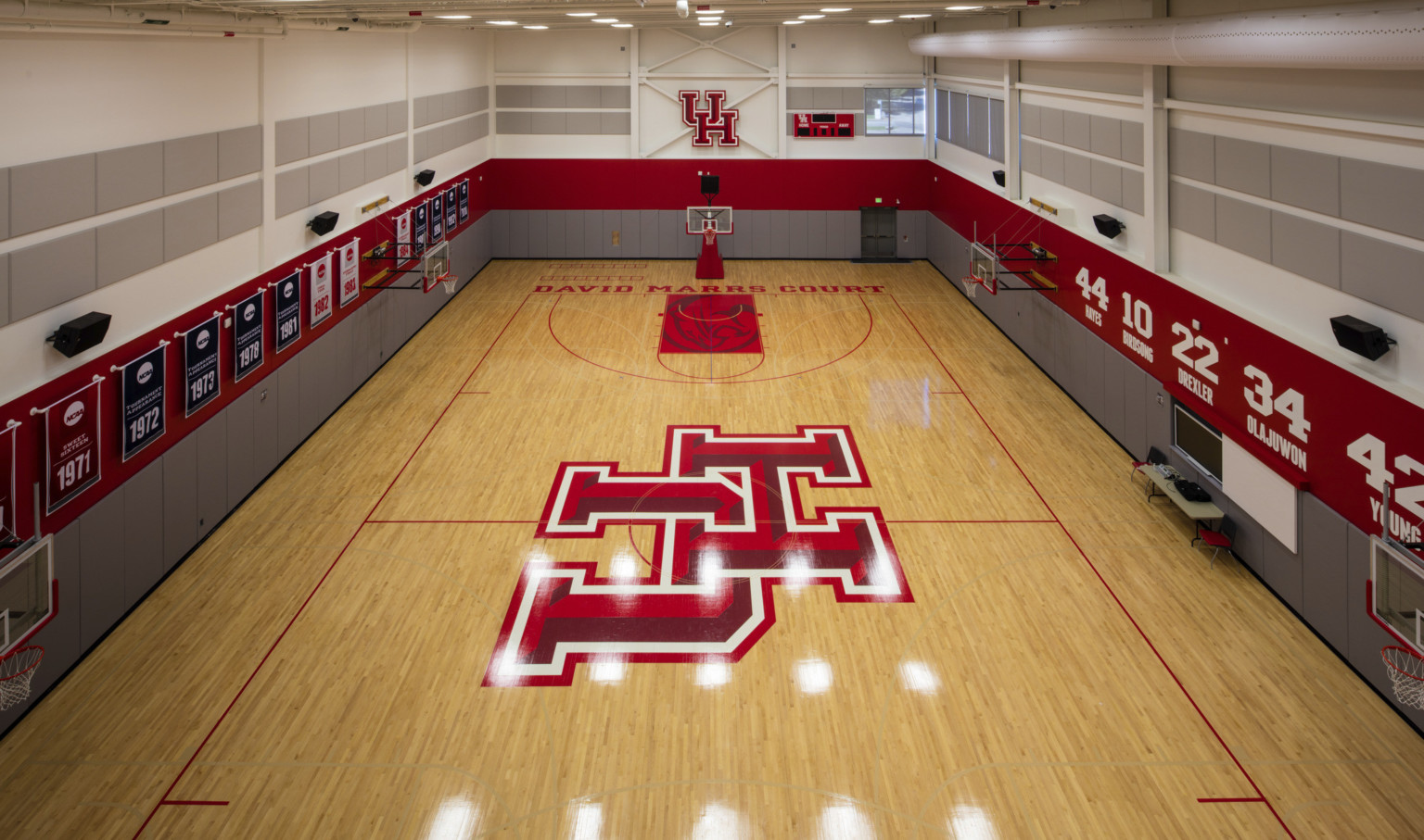 Wood practice court with basketball hoops along sides and UH logo at center. Grey and red stripes on walls with banners left Wood practice court with basketball hoops along sides and UH logo at center. Grey and red stripes on walls with banners left