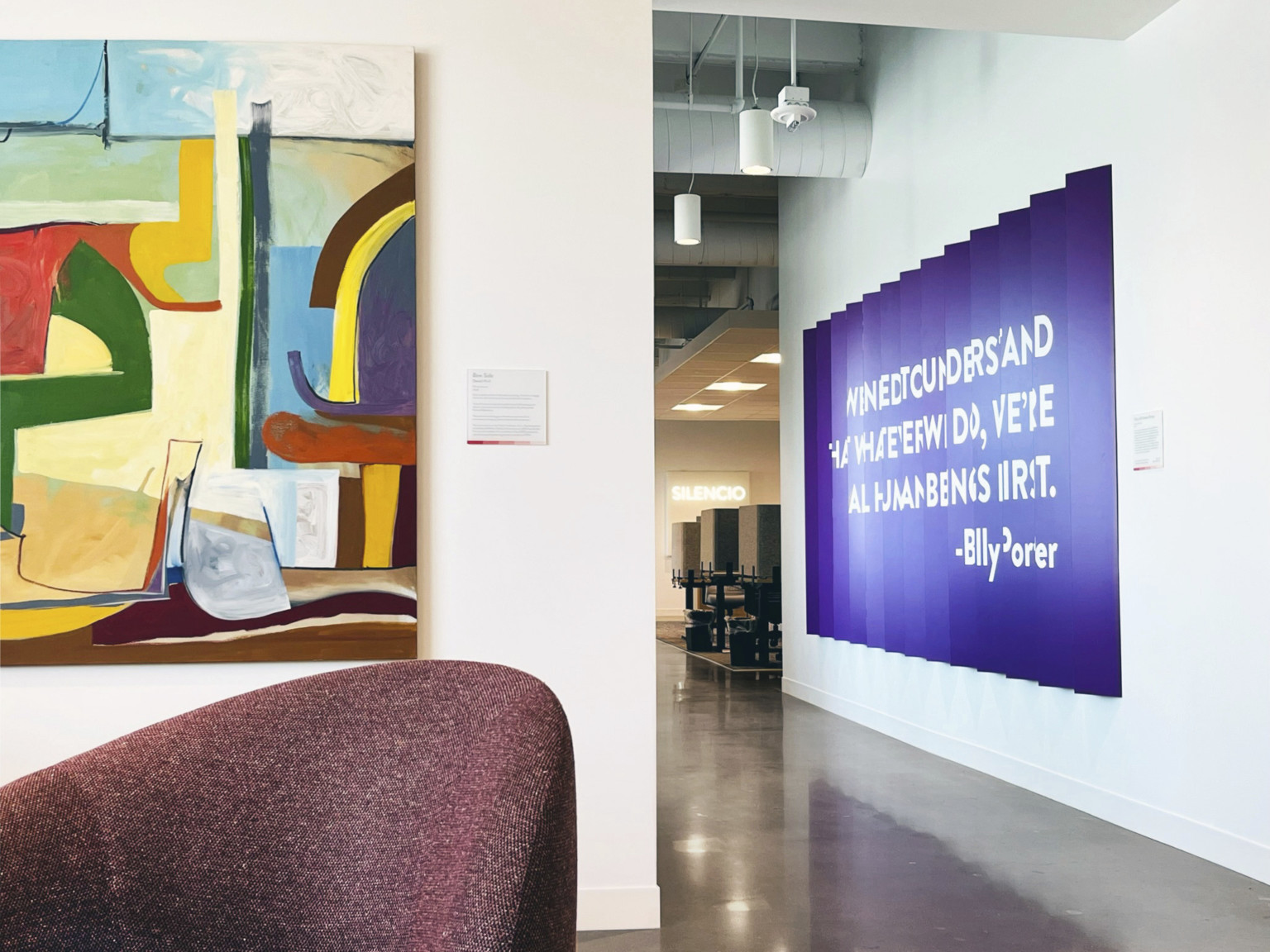 burgundy upholstered chair in front of colorful geometric painting and purple slat with white text installation on white walls