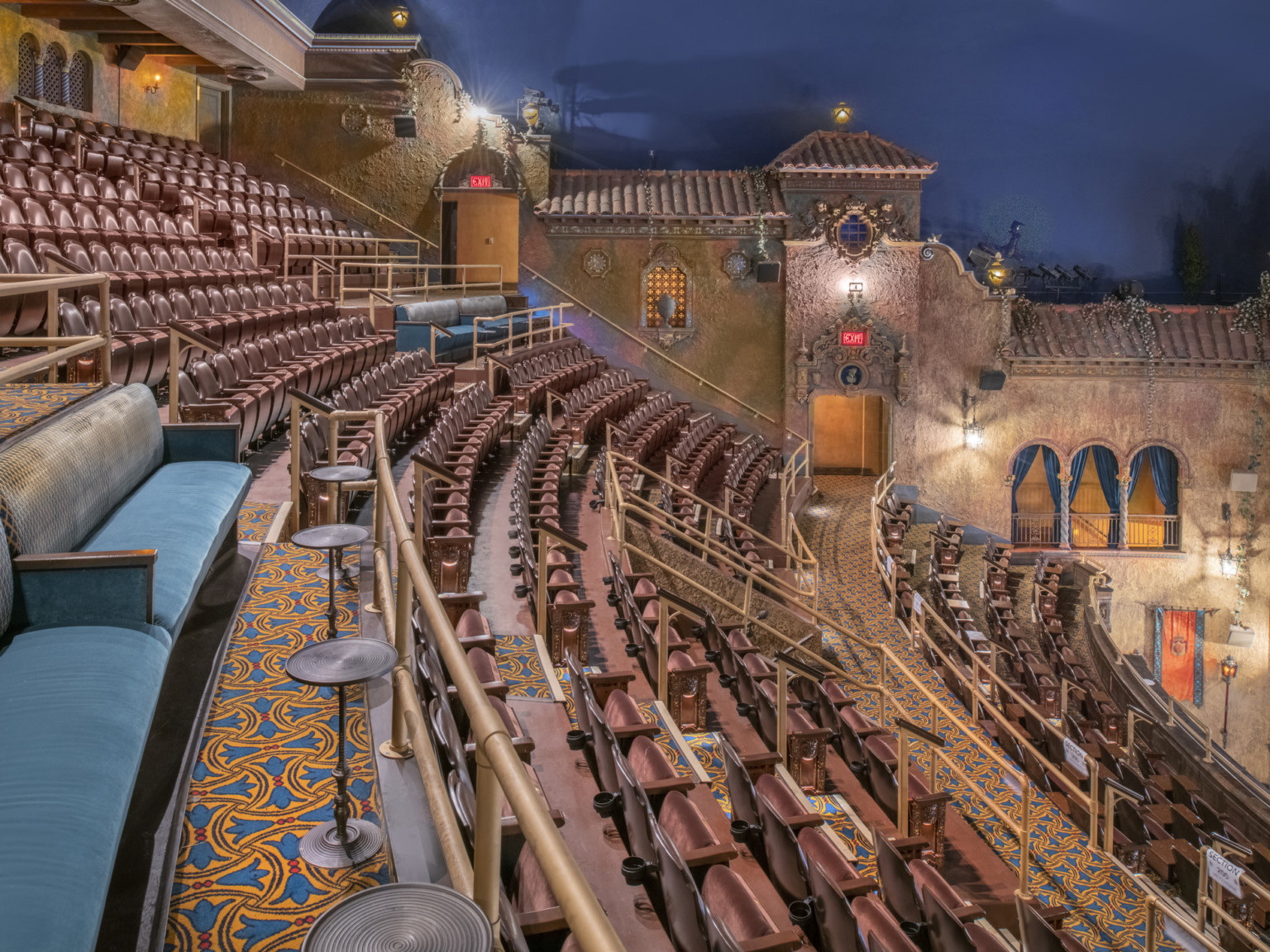 Theater balcony with mix of stadium and couch seating. Lined with faux building facade and illuminated windows, some curtained