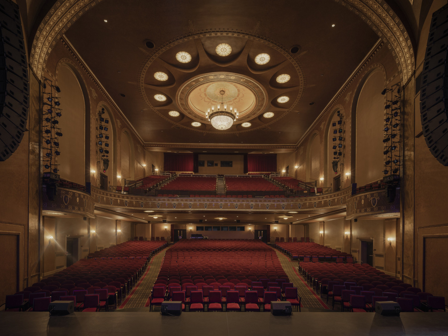 Theater from stage framed by curving proscenium arch looking to red audience seats, large balcony. Round light details encircling chandelier