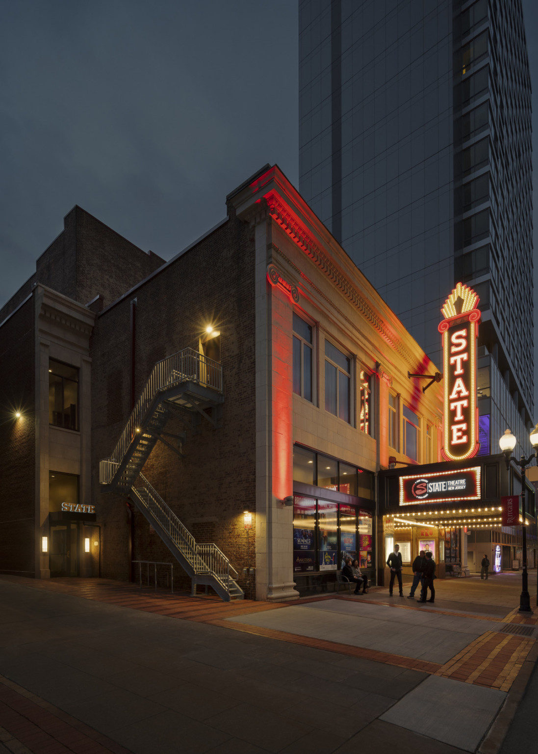 Theater exterior from corner with illuminated retro neon blade sign reading State over marquee. Stairs on side of building