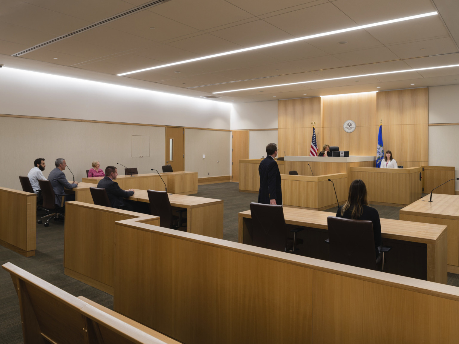 Interior courtroom with wood benches, rails and judge's seat. Wood panel accent wall at back with seal, recessed light rods