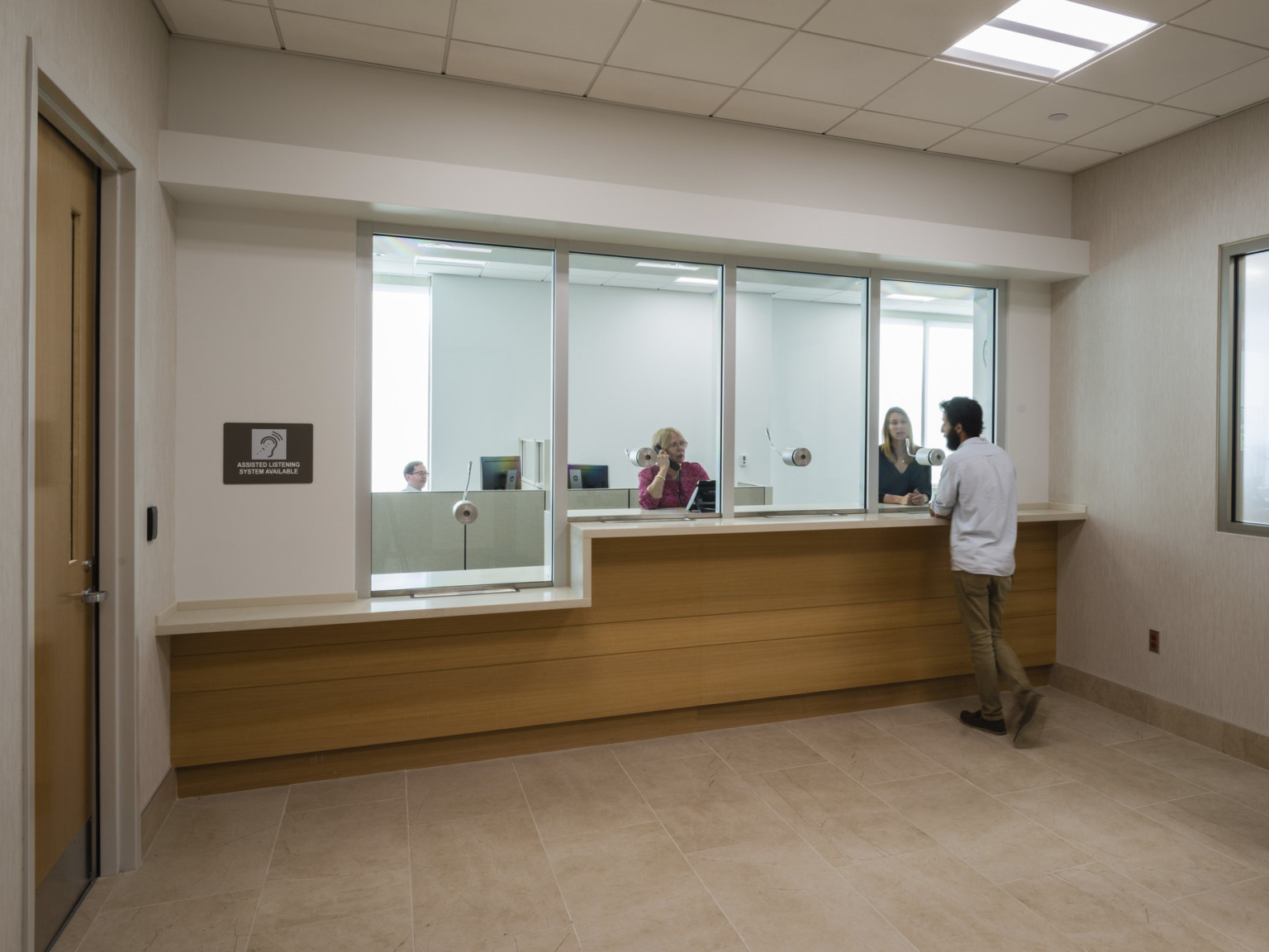 Interior reception area with wood panel desk and glass separating workers from public. White walls and tiled ceiling, wood door