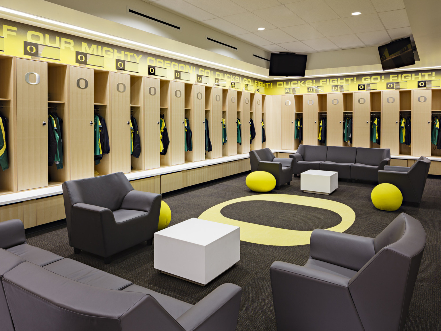 Locker room with yellow oregon logo center with wood lockers, black couches and arm chairs. UO fight chant along wall trim