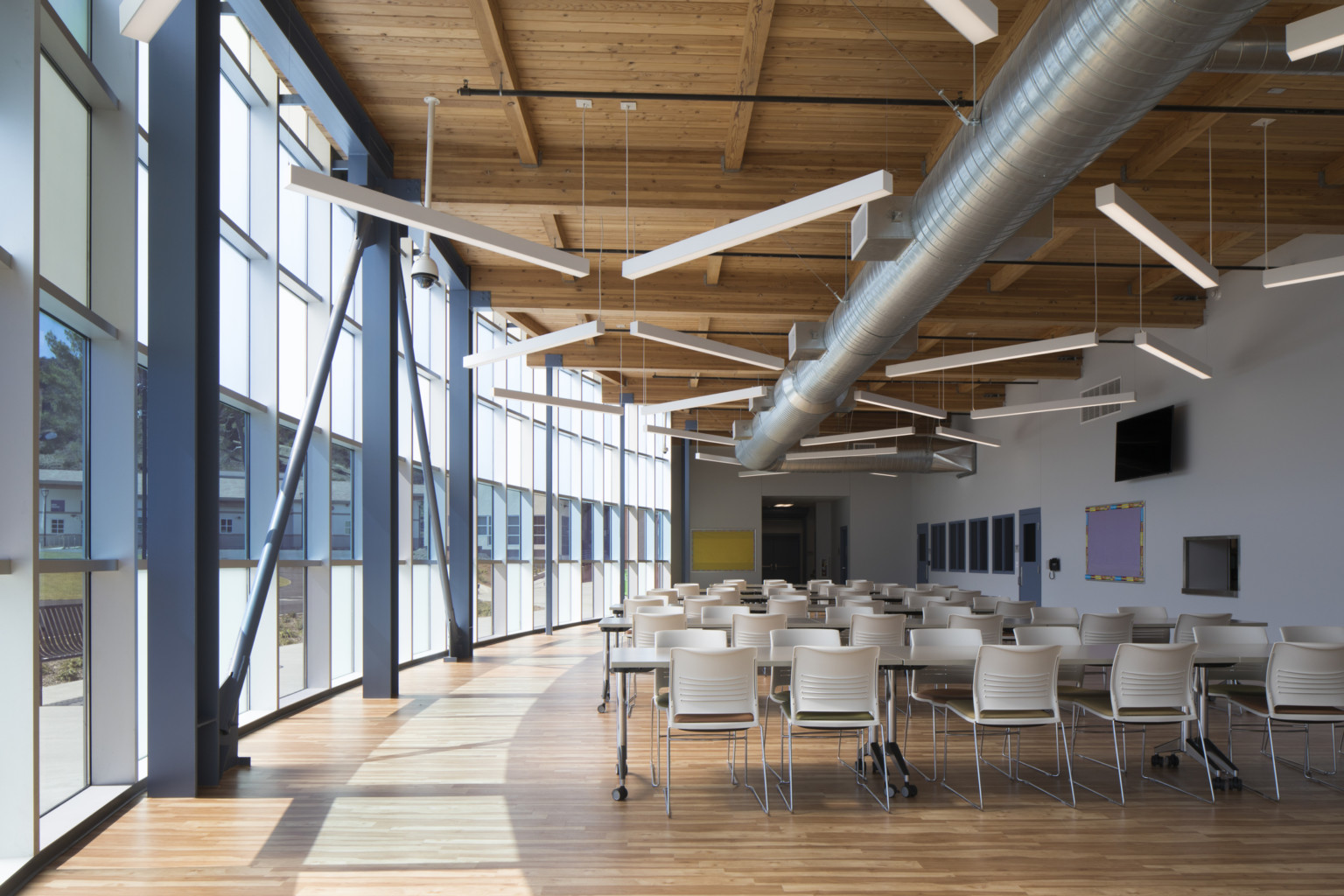 Interior view with curved glass wall, left. Right, a seating area with tables under wood ceiling with exposed beams and ducts