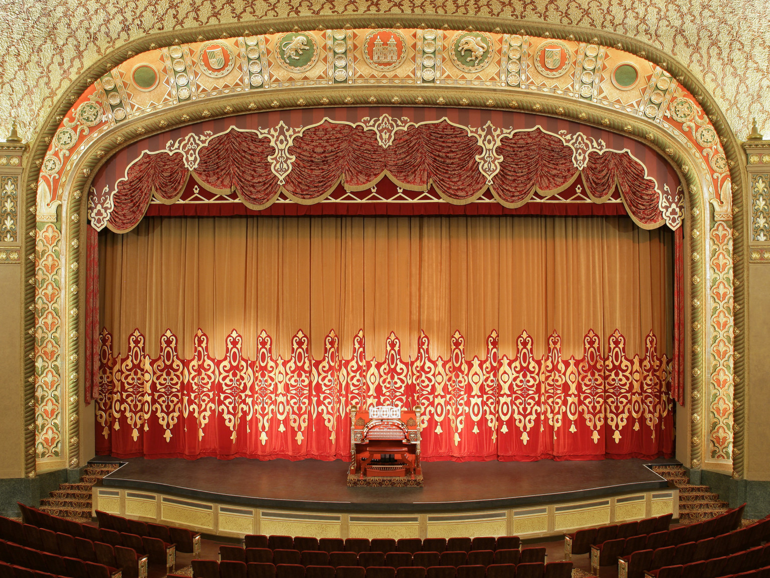 Stage with gold and red curtain and gold carved proscenium with green and orange accents a red and gold instrument at center