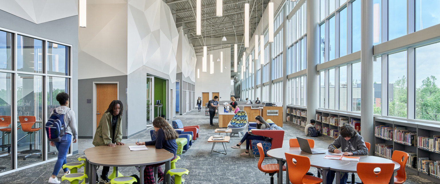 Hallway with double height windows, right, above low book shelves. Left, rooms under white organic sculptures. Seats between