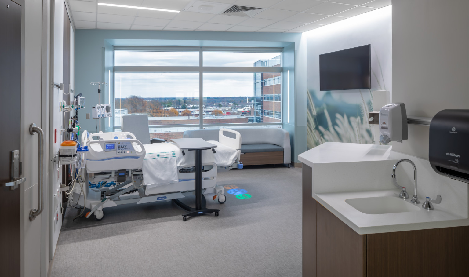 Hospital room with bed beside large window. Mural of grain on right wall with screen