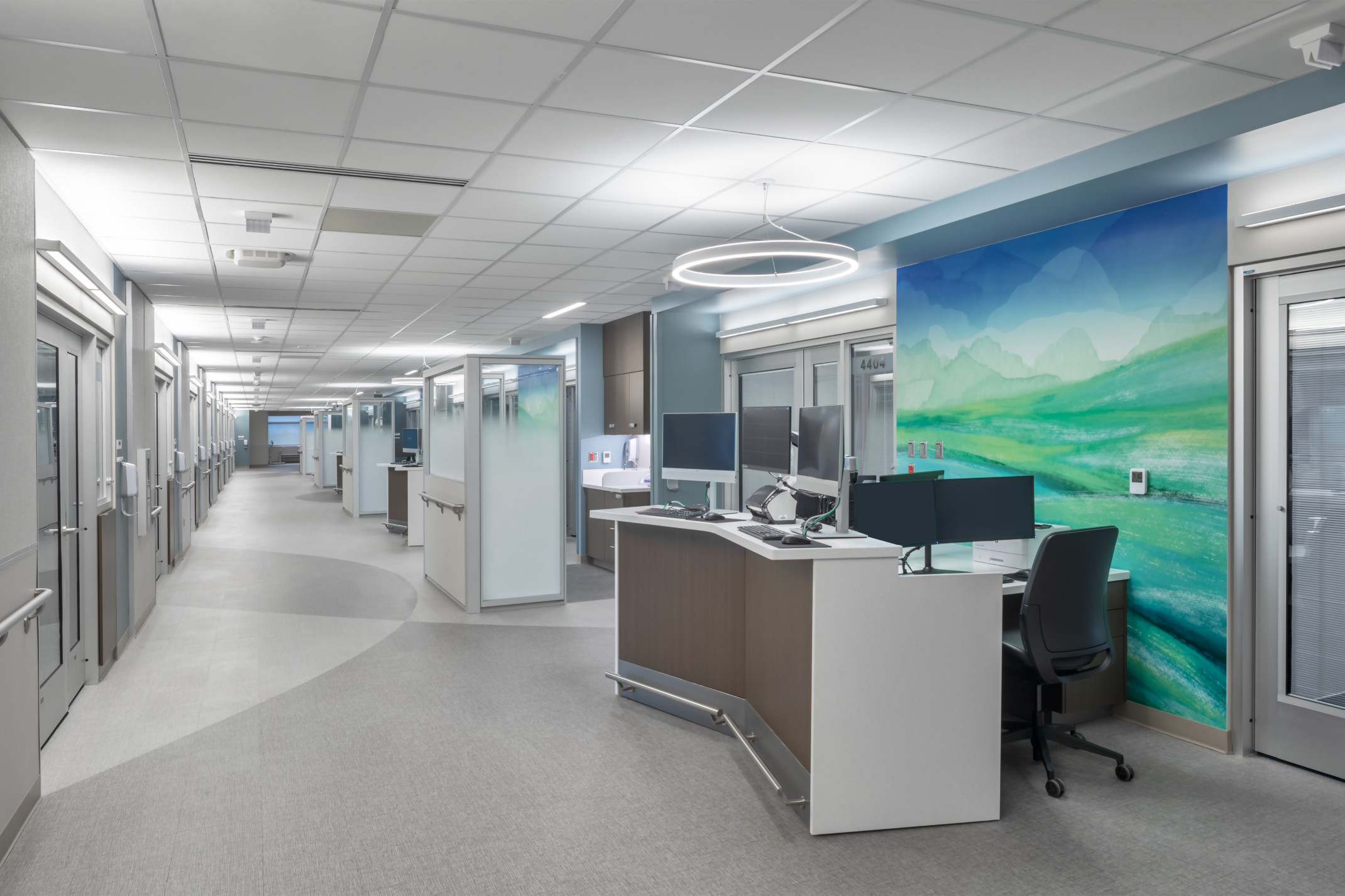 Hospital corridor with angled desk with multiple monitors in front of abstract blue and green mural. Glass partitions beyond Hospital corridor with angled desk with multiple monitors in front of abstract blue and green mural. Glass partitions beyond