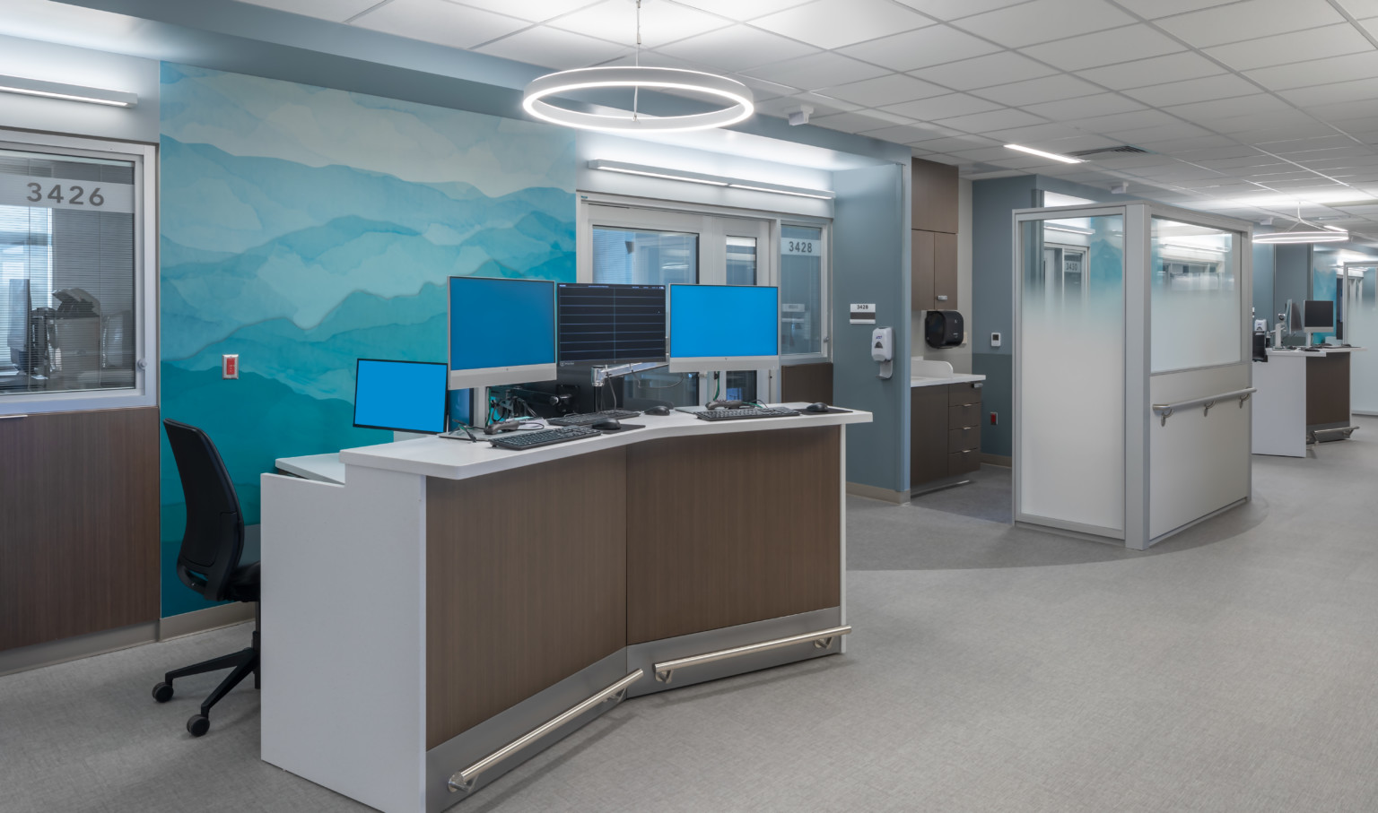Hospital corridor with angled desk with multiple monitors in front of abstract blue and green mural. Glass partitions beyond