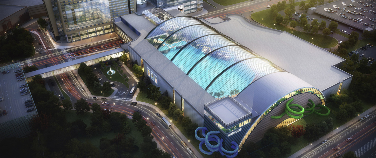 Aerial view of indoor waterpark with arching skylights over roof. Blue and green water slides curl out sides of grey building