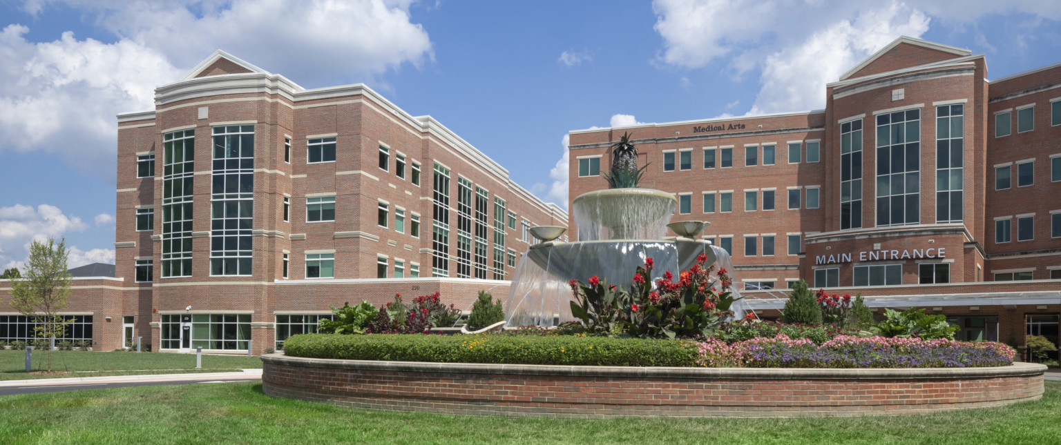 Exterior view of a brick hospital with central towers on each wing with windows down the center. Lawn to front with brick lined fountain